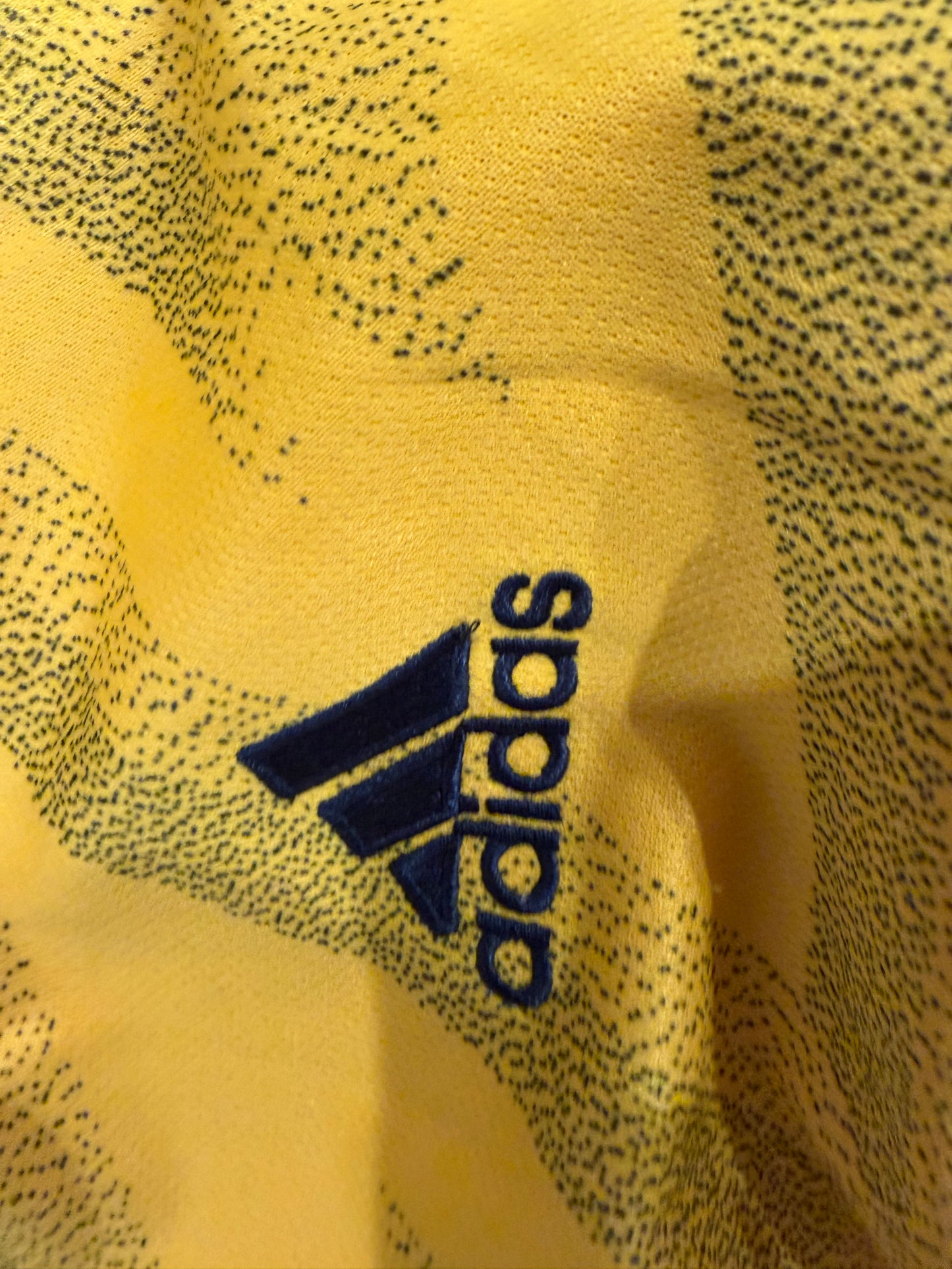 Close-up of a yellow Adidas garment with a dark navy blue logo and a fine dark speckled pattern across the fabric.