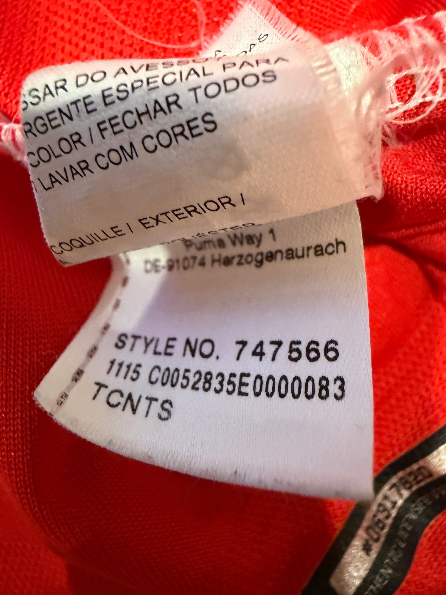 Close-up of a red garment's interior labels, showing a white sewn-in tag with black text including 'Puma Way 1' and 'STYLE NO. 74756', alongside a Portuguese care/label tag.