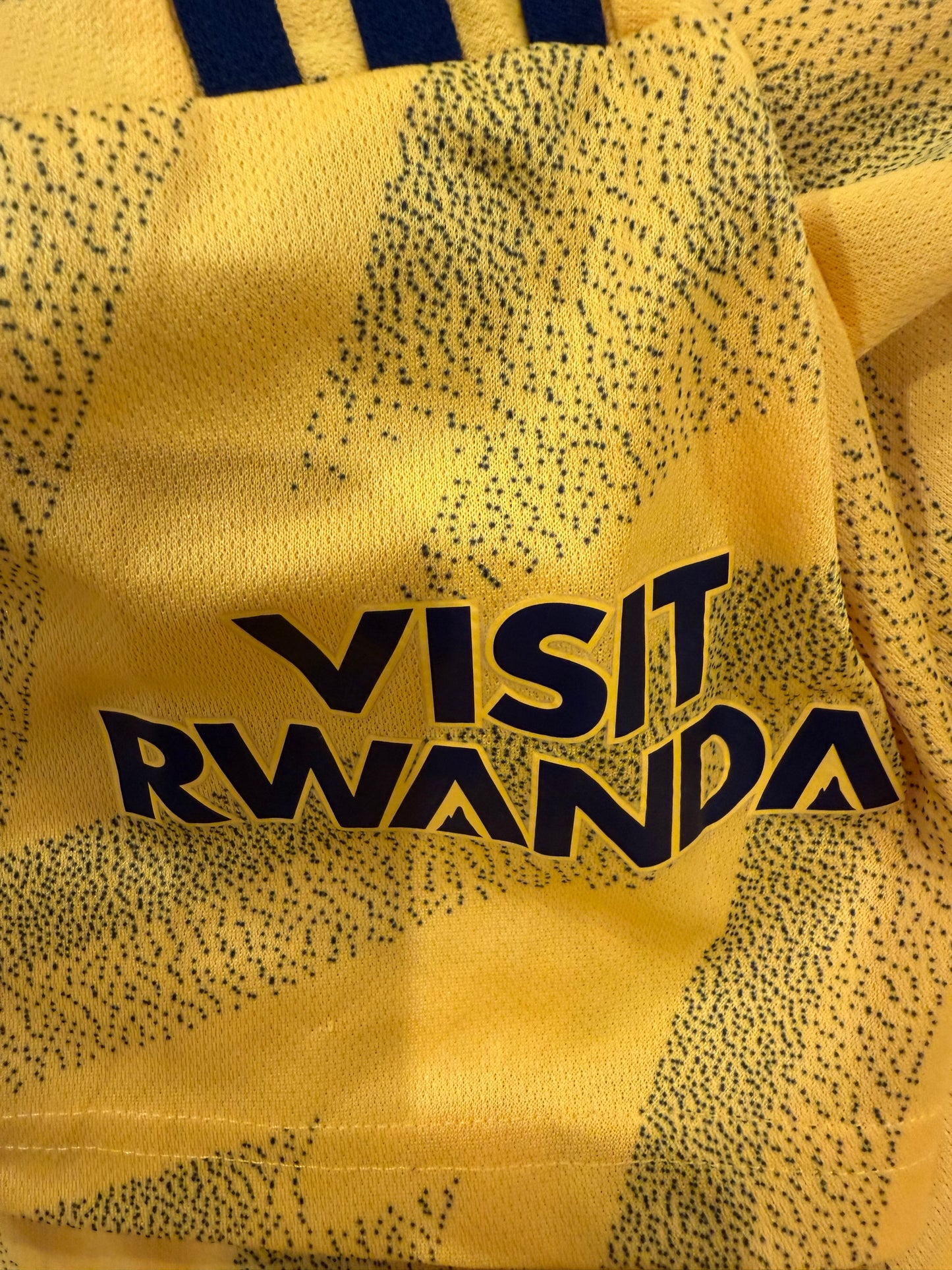 Close-up of a yellow sports jersey with a black dotted pattern, featuring the text 'VISIT RWANDA' in bold black letters.