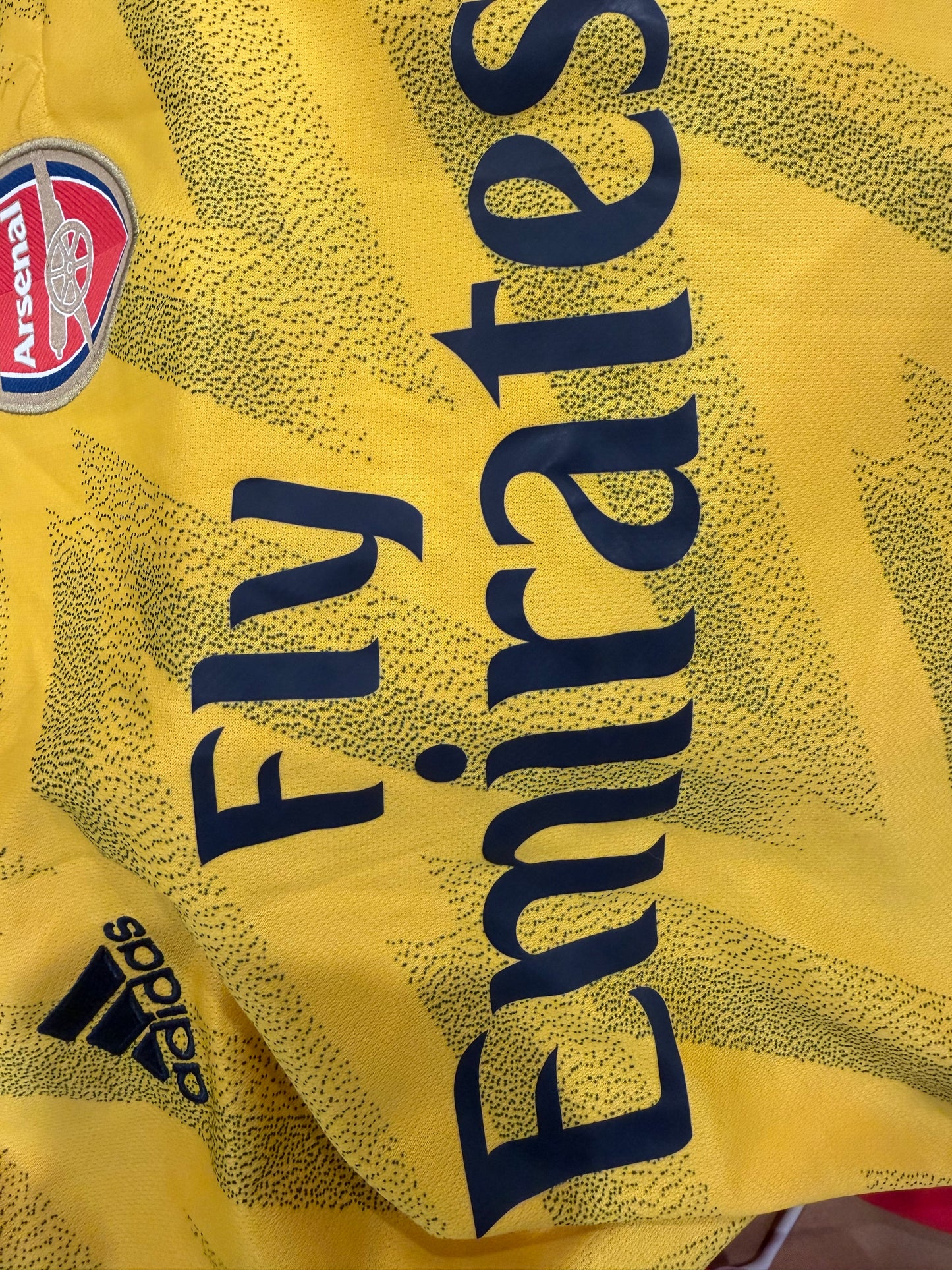 Close-up of a yellow Arsenal soccer jersey with dark blue 'Fly Emirates' sponsor text, the Arsenal club crest, and an Adidas logo.