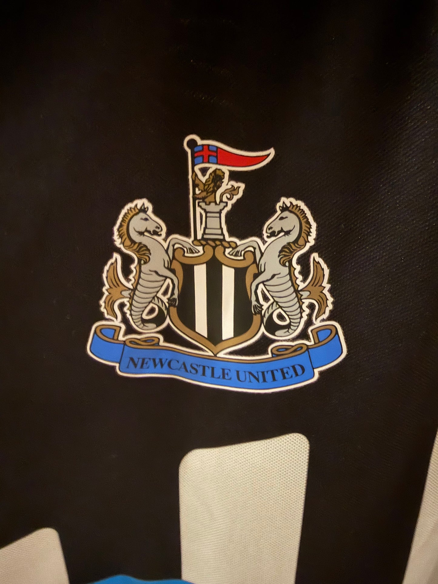 Close-up of a Newcastle United crest patch on a black garment, with part of a white sleeve visible below.