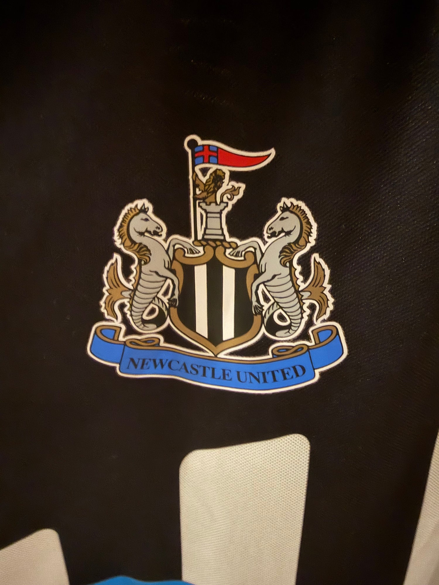Close-up of a Newcastle United crest patch on a black garment, with part of a white sleeve visible below.