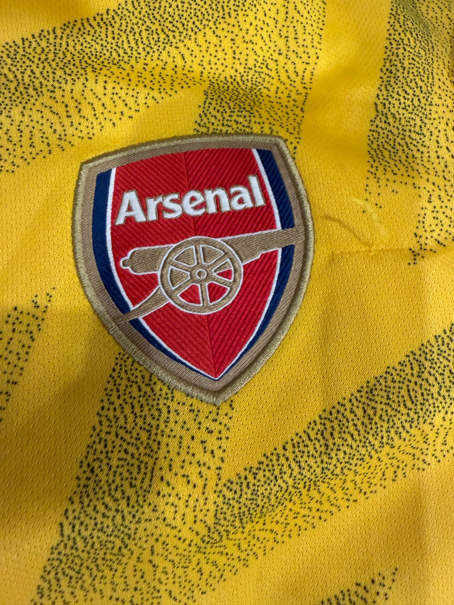 Close-up of an embroidered Arsenal football club crest patch on a yellow patterned jersey fabric.