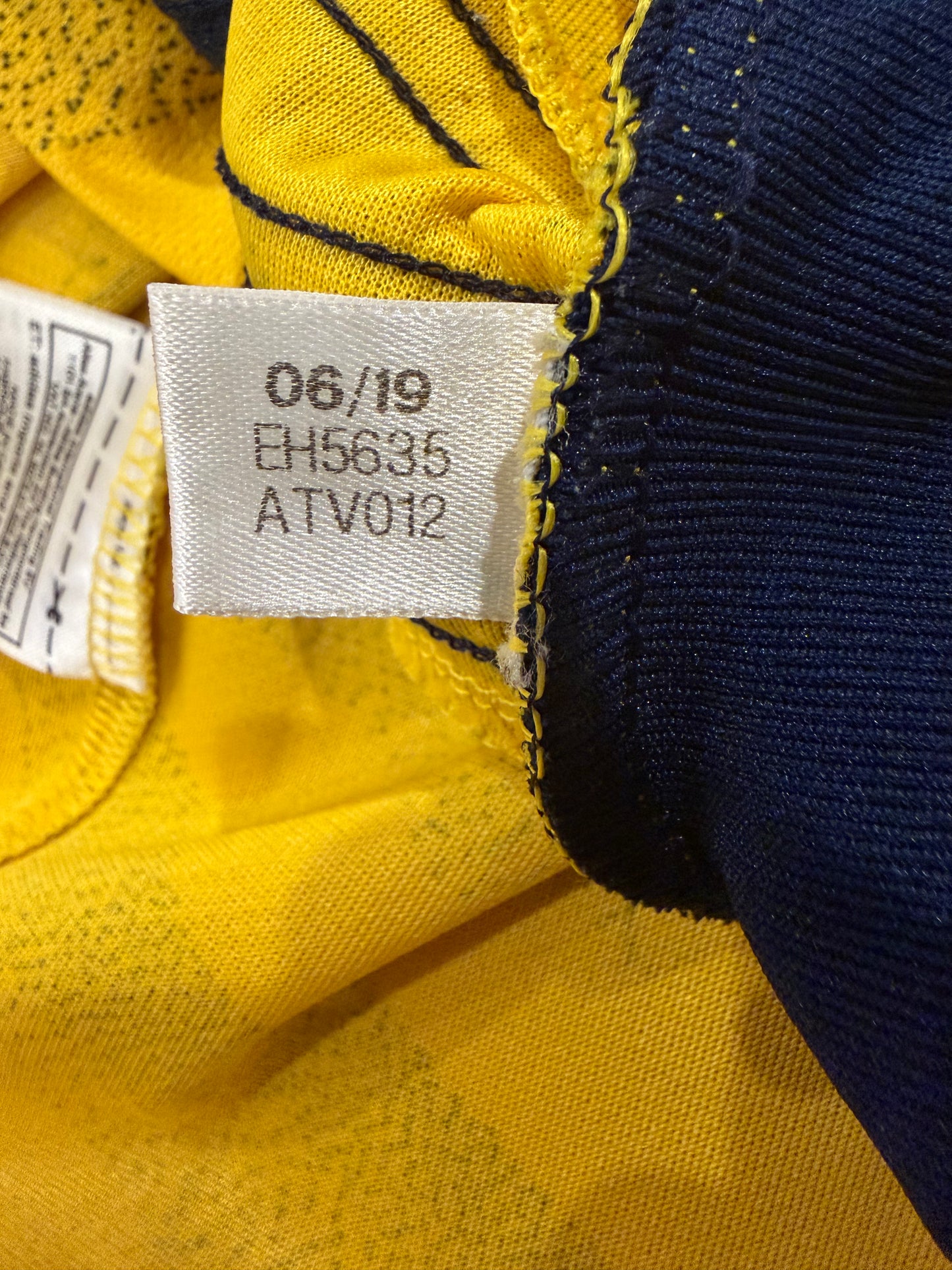 Close-up of a garment's inner neck area showing a white size tag with printed codes '06/19 EH5635 ATV012' stitched onto yellow and navy blue fabric.